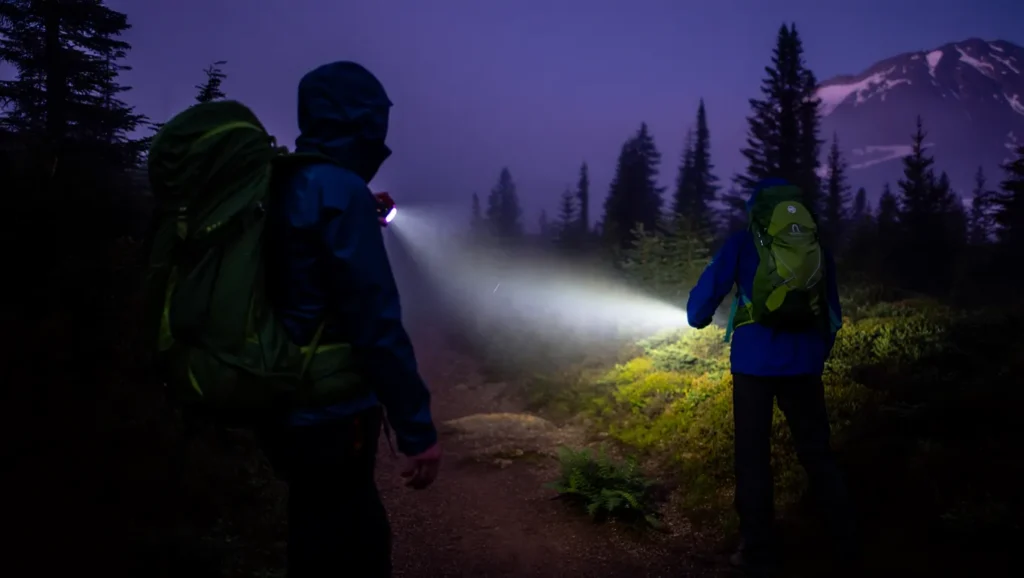 Person using a camping flashlight to navigate a trail at dusk, with beautiful landscape in background