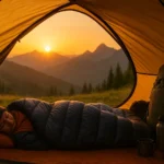 Premium ultralight down sleeping bag compressed into a small stuff sack next to a water bottle for size comparison, outdoors setting with mountains in background